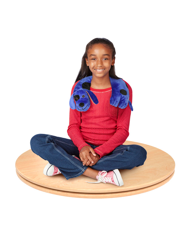 A child with dark skin tone and long black hair is sitting cross legged on the Spin Board. They are smiling and wearing a stuffed dog around their neck.