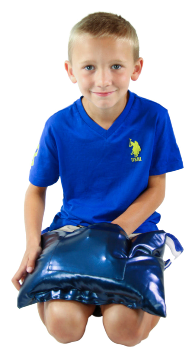 A child with light skin tone and short blonde hair is sitting on their knees with the Abilitations Vinyl Vibrating Weighted Lap Pad on their lap. Their left hand is in the pocket on the front of the pillow.