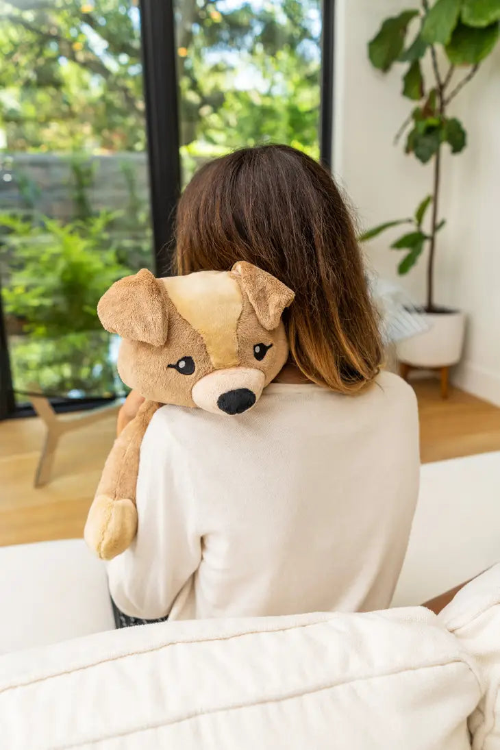 A person with long brown hair is sitting on a couch and holding the Dog Hugimals Weighted Stuff Animal over their left shoulder.