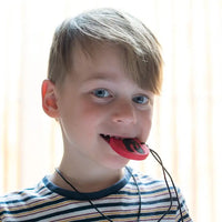 A child with light skin tone and short blonde hair has the red and black Dino Pendant in their mouth. They are holding it between their teeth and looking straight into the camera.