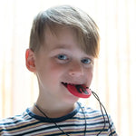 A child with light skin tone and short blonde hair has the red and black Dino Pendant in their mouth. They are holding it between their teeth and looking straight into the camera.