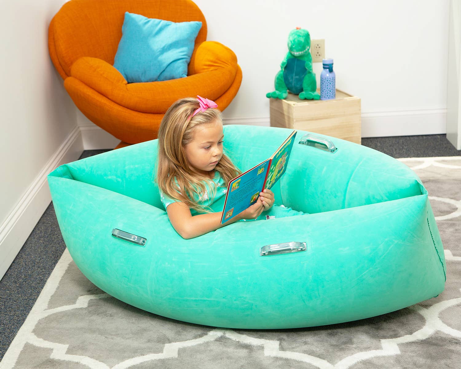 A child with light skin tone and long blonde hair is sitting in a light blue Peapod. They are leaning to the side and reading a book.
