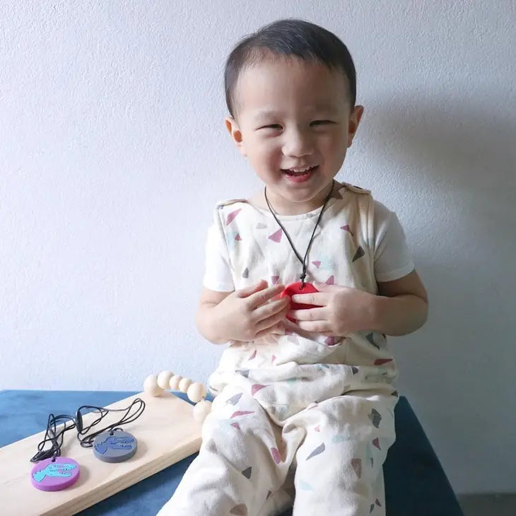 A child with light skin tone and dark brown hair is sitting on a piece of indistinguishable furniture. They are holding the red and black Dino Pendant and smiling, and a board with the other color variants of the pendant are next to them.
