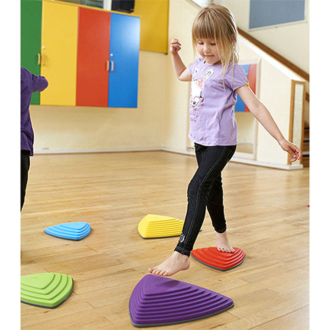 A child with light skin tone and long blonde hair moves their left foot towards a large purple Gonge Riverstone while their right leg balances on a small red one.