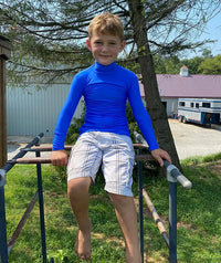A child with light skin tone and short blonde hair is sitting on a piece of play equipment outside. They are smiling and wearing a blue Mock Turtleneck Long Sleeve Compression Shirt.