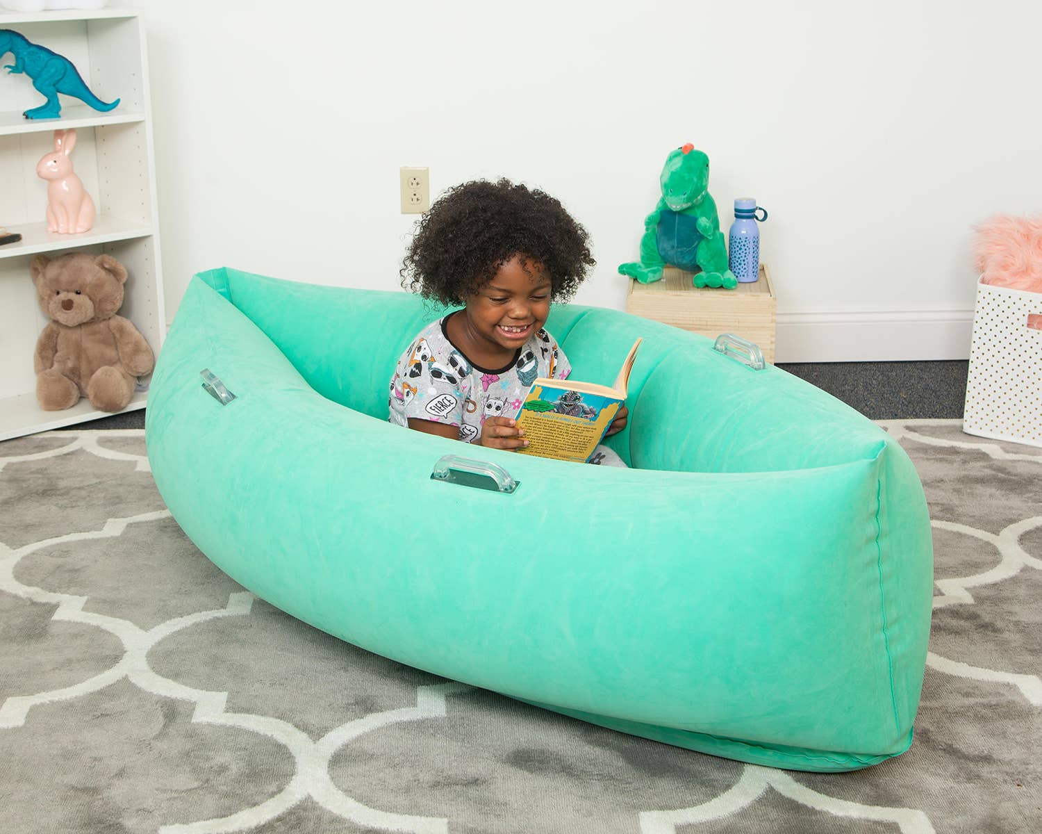 A child with dark skin tone and chin length curly black hair is smiling and holding open a book. They are sitting in a Teal Hugging Peapood (60"),