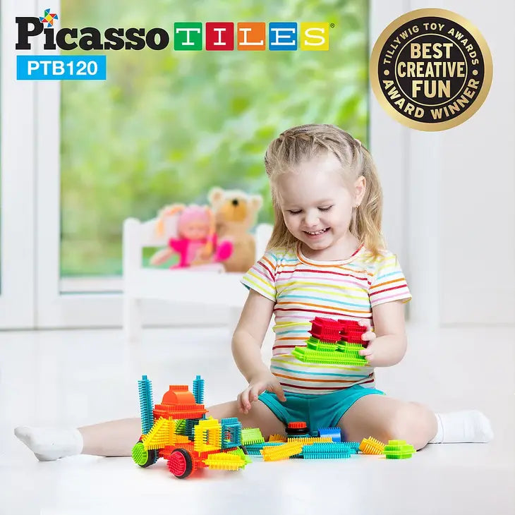 A child with light skin tone and shoulder length blonde hair sits on the floor with one leg splayed out in front of them. They are holding several BristleLock Building Blocks and there is a small vehicle built with the same blocks sitting in front of them.