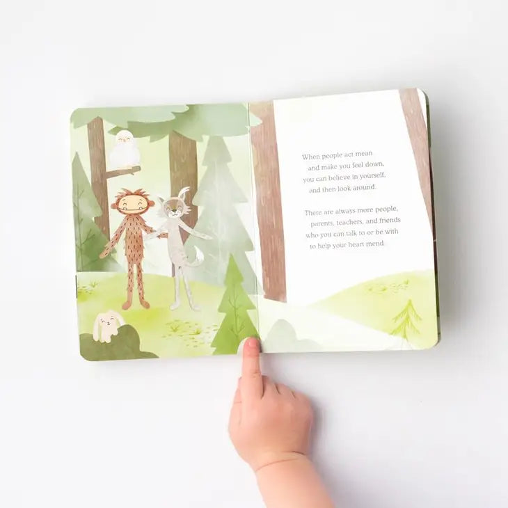 A tiny hand with light skin tone puts its finger in the middle of the open board book that comes with Slumberkins Maple Bigfoot Snuggler.
