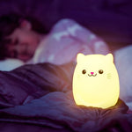 A Cat LED Night Light Bluetooth Speaker is lit up in the foreground. Behind it is a child under the covers.