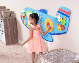 A child with medium skin tone and two high black braids stands next to the Airplane Activity Wall Panel in a bedroom. They are touching the clock panel and appear to be in a bedroom.