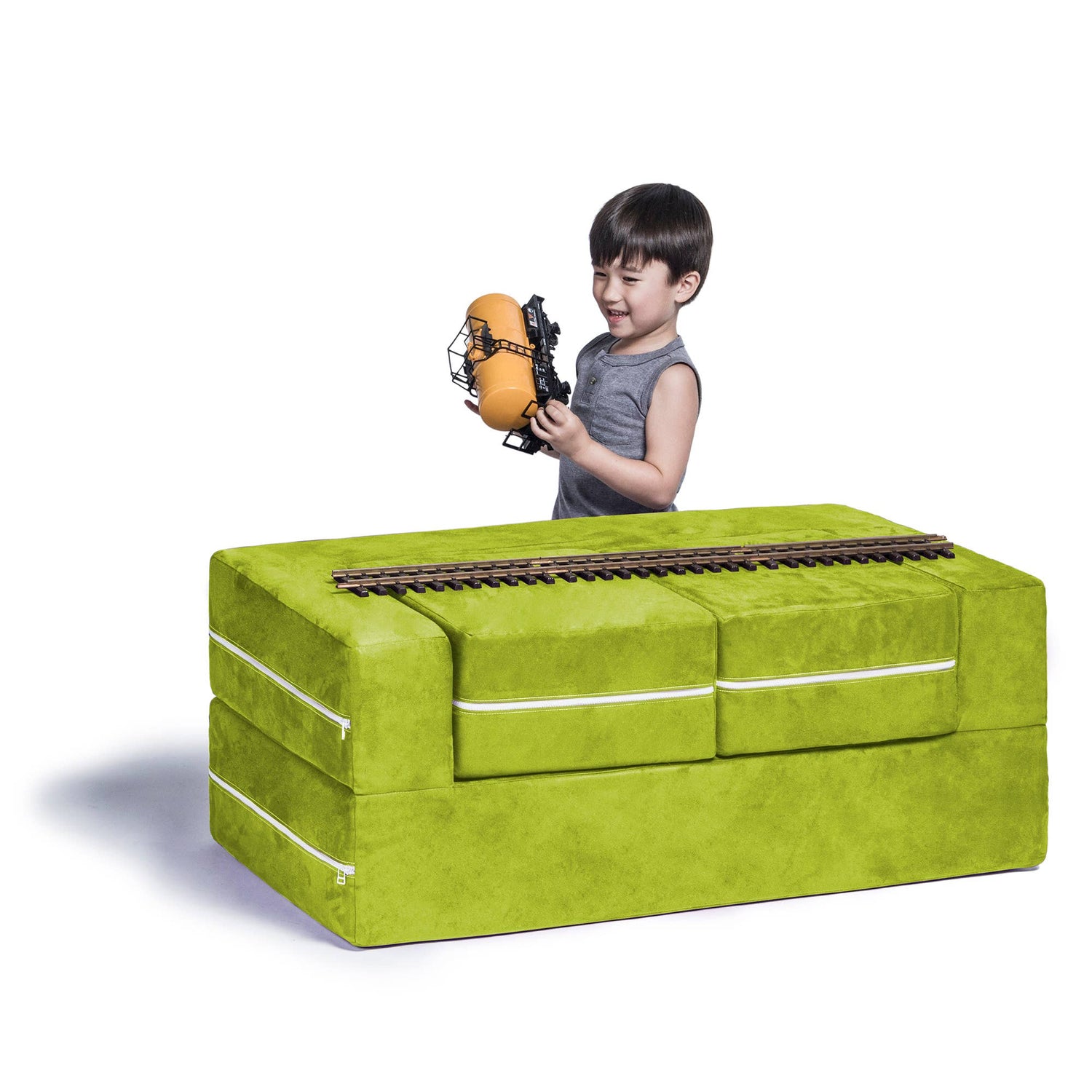 A child with light skin tone and short black hair is standing on the far side of the Jaxx Zipline Modular Kids Loveseat and Ottomans. They are holding a toy train piece, and there is a set of tracks running scross the length of the loveseat.