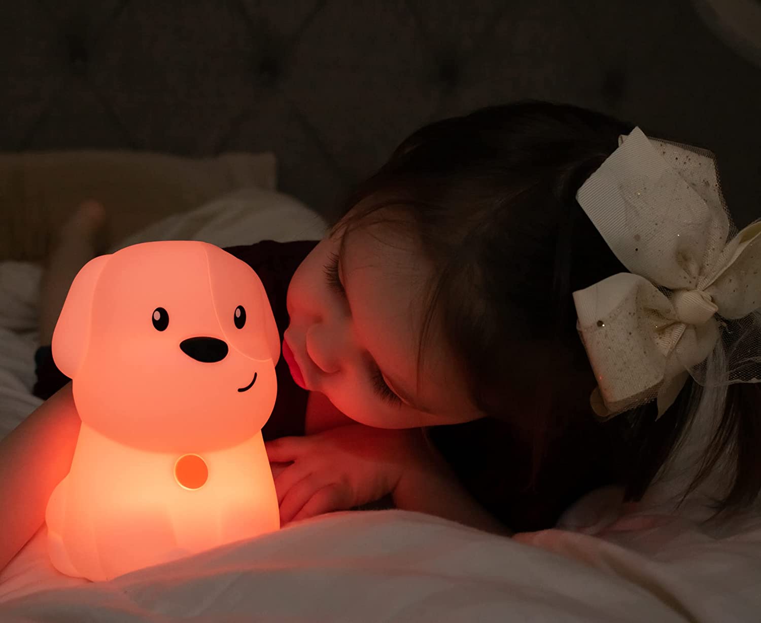 A child with light skin tone and long brown hair makes a face at the Dog LED Night Light Bluetooth Speaker. It is dark in the room and the Dog is lit up.