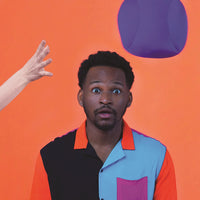 A person with dark skin tone and short black hair has a surprised facial expression. They are wearing a colorful shirt against a orange backdrop. There is an isolated hand with light skin tone that appears to be throwing a purple Dohzee over their head.