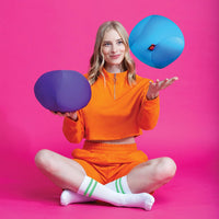A person with light skin tone and long blonde hair sits with their legs crossed. They are wearing a bright orange jump suit against a bright pink background. They are holding a purple Dohzee in one hand and appear to be tossing a blue Dohzee with the other.