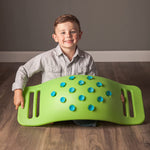 A child with light skin tone and short brown hair is kneeling on the floor and holding a green Teeter Popper on its side. The suction cups are displayed.