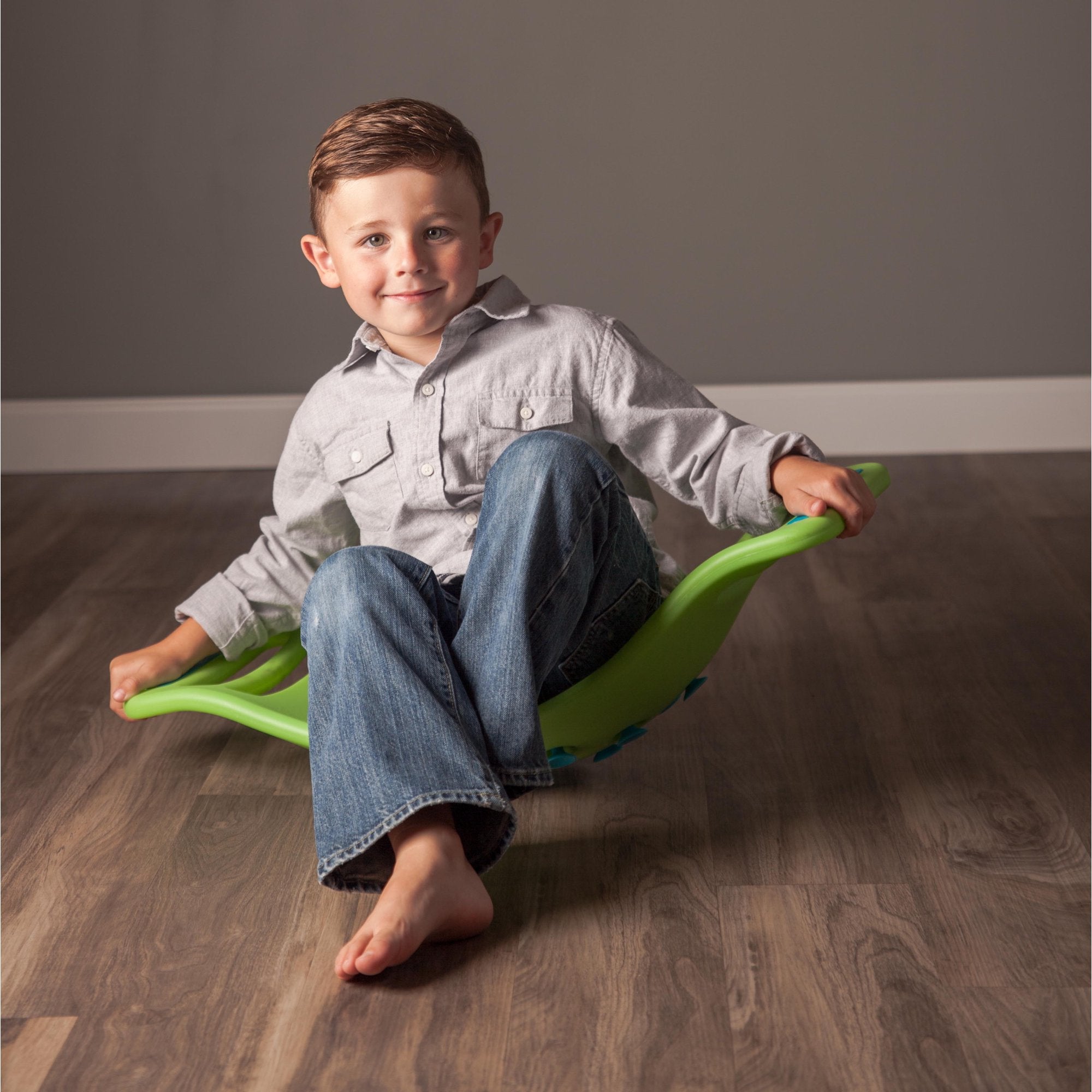 A child with light skin tone and short brown hair is sitting in the green Teeter Popper. Their legs are crossed and they are leaning back.