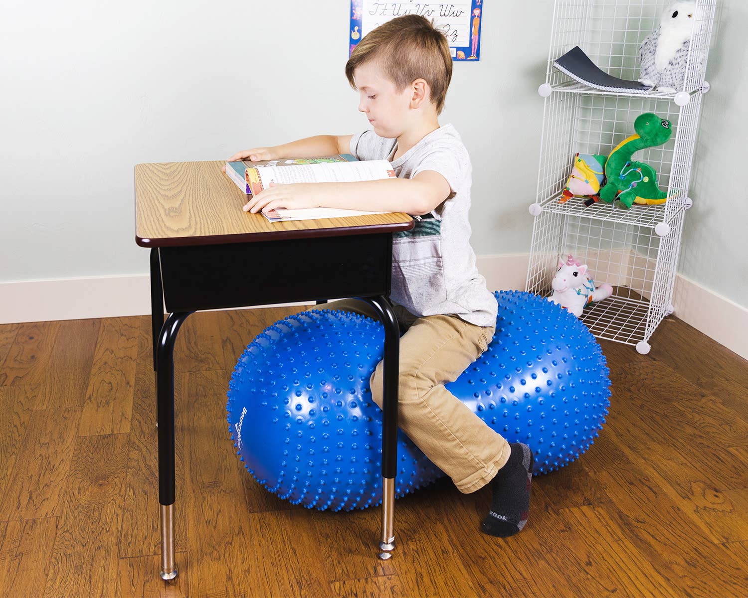 A child with light skin tone and short brown hair sits on a blue Peanut Stability Ball. They are at a desk with a book open.