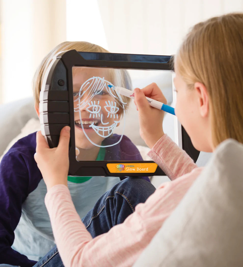 A child with light skin tone and long blonde hair holds up a Glow Board in front of another child with light skin tone. The first child is sketching the face of the other onto the Glow Board with a marker.