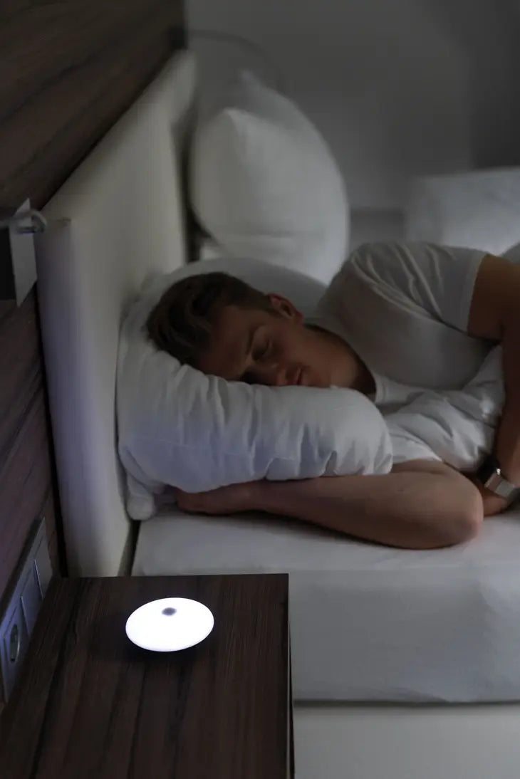 A person with light skin tone and short blonde hair is in bed facing an end table in the foreground.  Their eyes are closed and one hand is positioned under the pillow. There is a white light emitting from the Troika USB Rechargeable Meeting Timer.