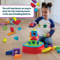 A child with medium skin tone and two brown pigtails kneels behind the platform and raises a red foam piece above a small structure. The text reads: The soft foam blocks are easy to stack into tall, teetering towers on the special building platforms.