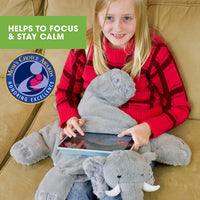 A child with light skin tone and long blonde hair sits on a couch. They hae the Barmy Weighted Lap Elephant on their lap and an Ipad resting on top of the elephant.