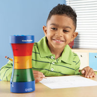 A child with dark skin tone sits at a desk with a piece of paper on it. They are looking at the Time Timer Visual Timer and Clock and smiling.
