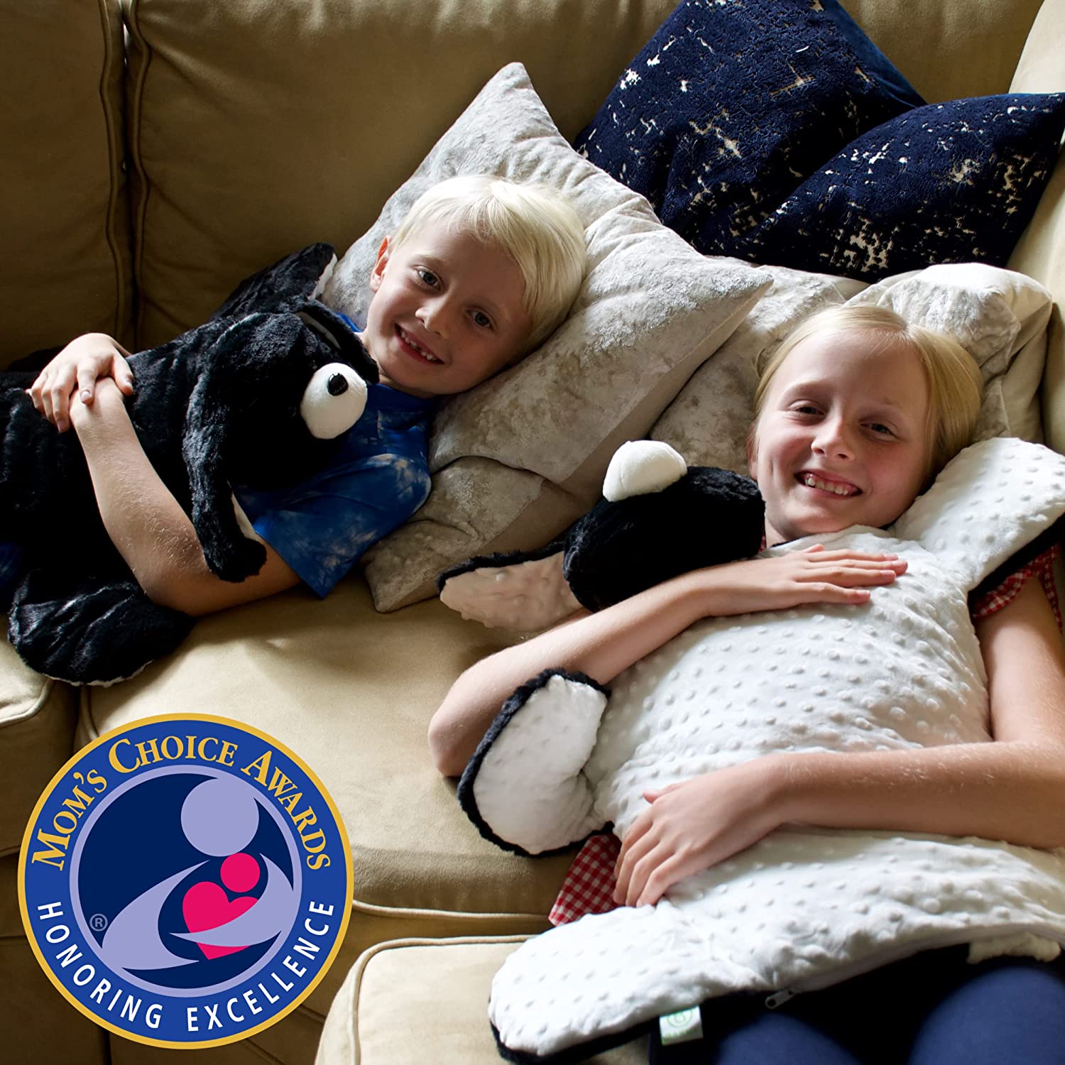 Two children lie on a couch with their heads close together. One has light skin tone and short blone hair; the Barmy Weighted Lap Dog is on their chest and their arms are crossed over the top. The other child has light skin tone and long blonde hair and the Barmy Weighted Lap Dog is upsidedown on their chest, revealing the textured minky belly of the dog.