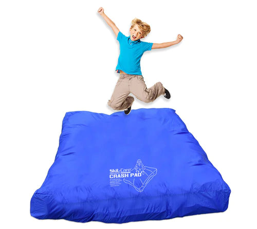 A child with light skin tone and short blonde hair is jumping with their knees up and arms stretched out over a Crash Pad.