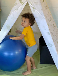 A child with medium light skin tone and short, brown, curly hair is standing next to an exercise ball. They are wearing a pair of gray Unisex Sensory Compression Shorts.
