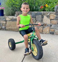 A child with medium light skin tone and very short brown hair is sitting on a tricycle and smiling. They are wearing a lime green sleeveless sensory compression shirt and black shorts.