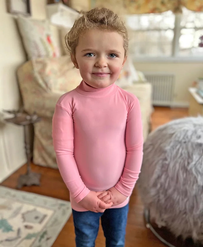 A child with light skin tone and blonde hair that is pulled back is standing with their hands clapsed. They are wearing a pink Mock Turtleneck Long Sleeve Compression Shirt.