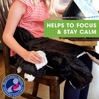 A child with light skin tone and long blonde hair is sitting on a wooden chair at a kitchen table. They have the Barmy Weighted Lap Dog on their lap, and they are holding up the long ear. The text reads: Helps to focus and stay calm.