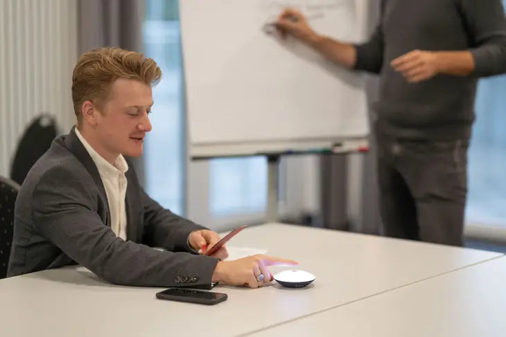 A person with light skin tone and short blonde hair is wearing a suit and sitting at a large table with one finger perched over a Troika USB Rechargeable Meeting Timer. In the background someone is standing in front of a white board and writing on it.