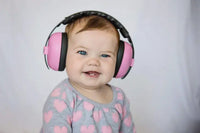 A baby with light skin tone and short brown hair is wearing the Pink Petal Baby Earmuffs. They have blue eyes and are smiling, revealing two bottom teeth.