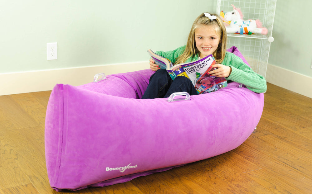 A child with light skin tone and long blonde hair is smiling and holdig an open workbook. They are sitting in a purple Hugging Peapod (60").