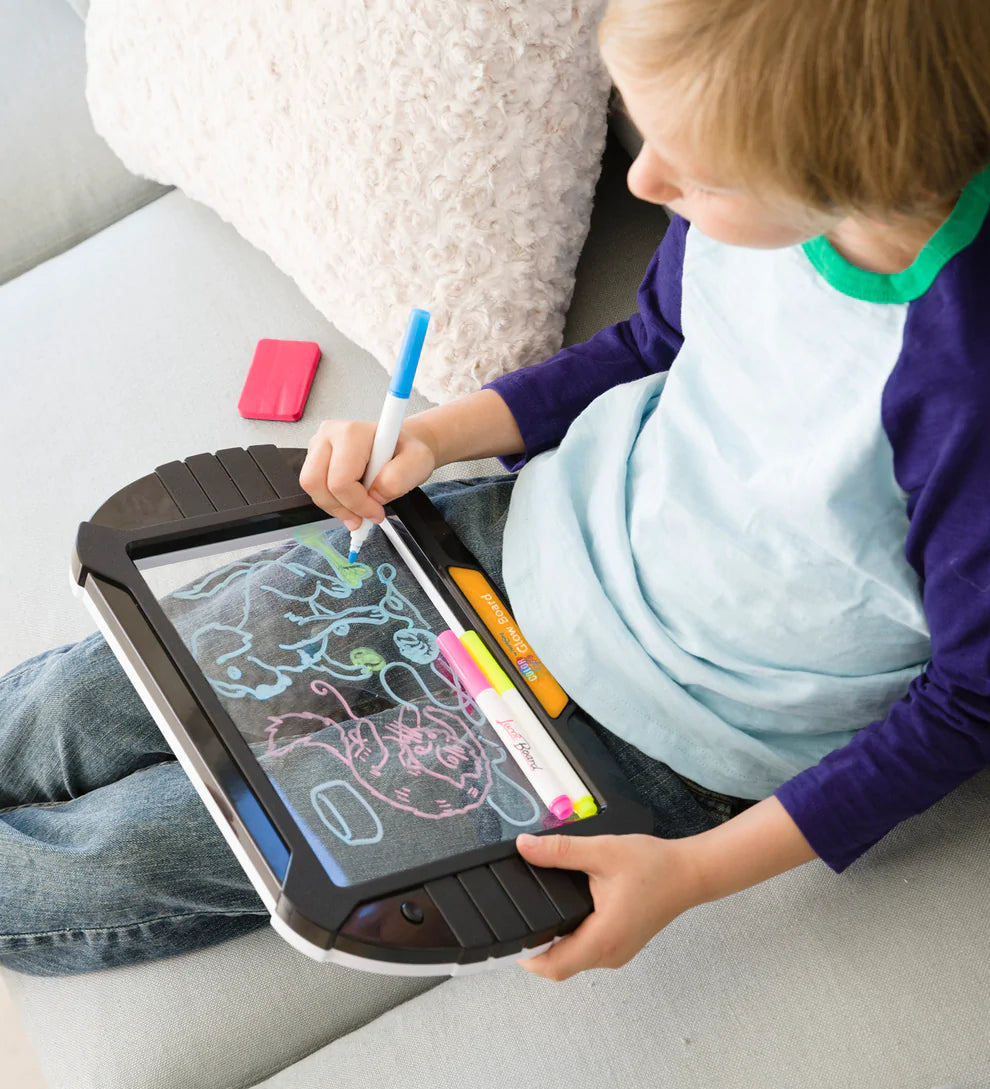 A child with light skin tone and short blonde hair sits on a couch, drawing on the Glow Board.