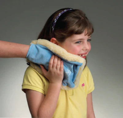A child with light skin tone and shoulder length brown hair holds the Vibrator Mitt up to their neck and face. There is an adult's hand in the mitt, and the child is smiling.