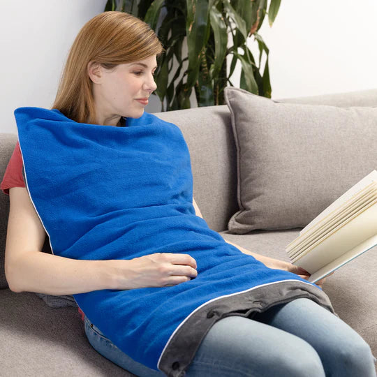 A person with light skin tone and long brown hair is sitting up on a couch with the Ms Bliss Aroma Blanket on their torso and upper legs. They are holding up an open book.