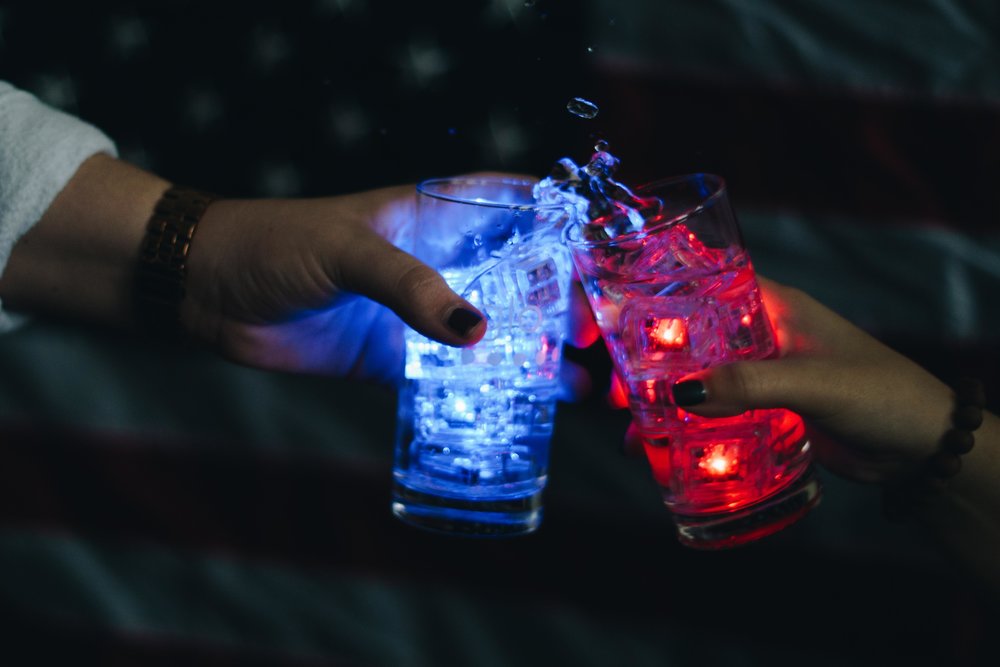 Two hands wearing black nail polish cheers two drinks with blue and red Glo Liquid-Activated Cubes in a dark setting.