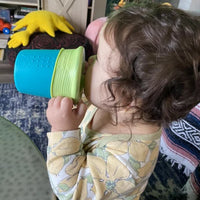 A toddler with light skin tone and brown curly hair is holding up the green and blue Universal Sippy Cup to their mouth and drinking from it.