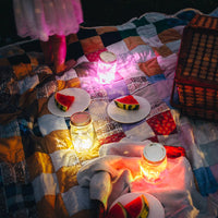 A picnic blanket is spread out in a yard at night. Several plates have cut up sllices of watermelon and a picnic basket is in the corner of the blanket. A child with a white dress stands on the blanket with only their lower half visible in the picture. There are three jars emitting a colorful glow, each containing a different color Glo Pal.