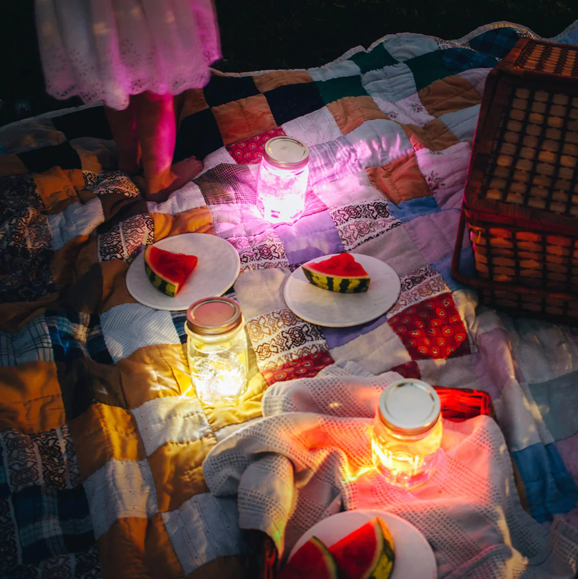 There is a picnic spread on a blanket in a dark yard. There are slices of watermelon on plates next to a picnic basket, and there are three mason jars with lids emitting three different colors from the Glo Liquid-Activated Cubes. There is a child wearing a white dress standing only half in the frame in the top left corner.