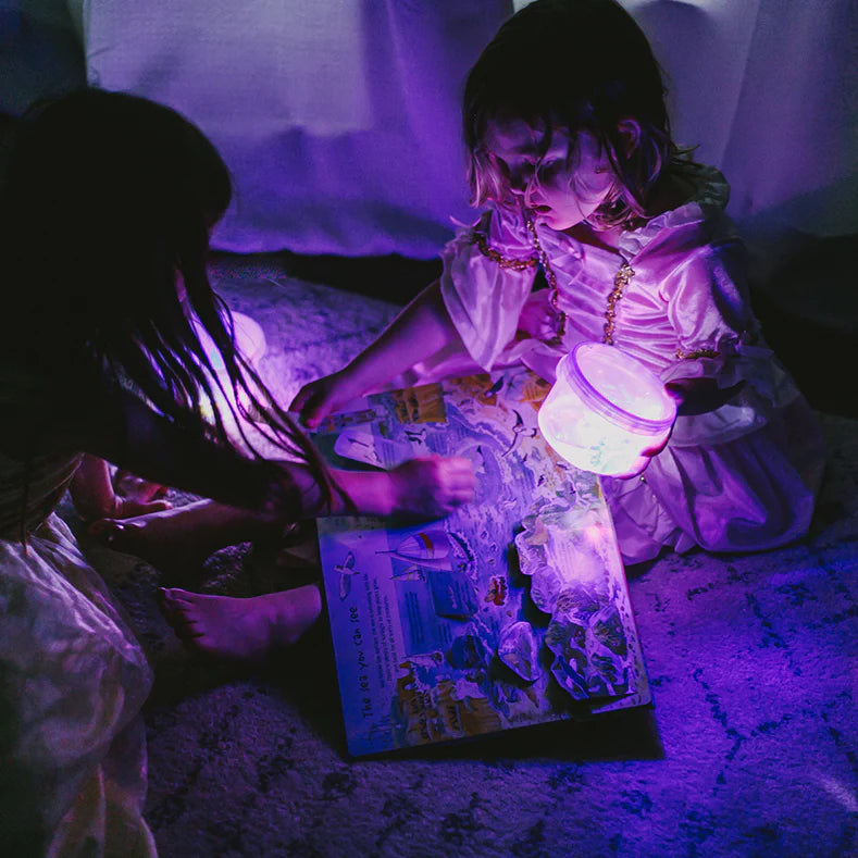 Two children sit in a dark room and a pop-up book is open on one of their laps. The child holding the book is also holding a clear jar being illulminated by a Glo Pals cube.