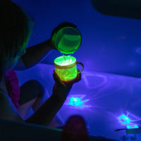 A child sits in a bathtub in a swimsuit. The bathroom is dark and the child is holding up two cups, pouring water from one to the other. The cup being poured into has a lit up Glo Pal cube, and the bathtub is illuminated with two Glo Pals cubes and a blue Character.