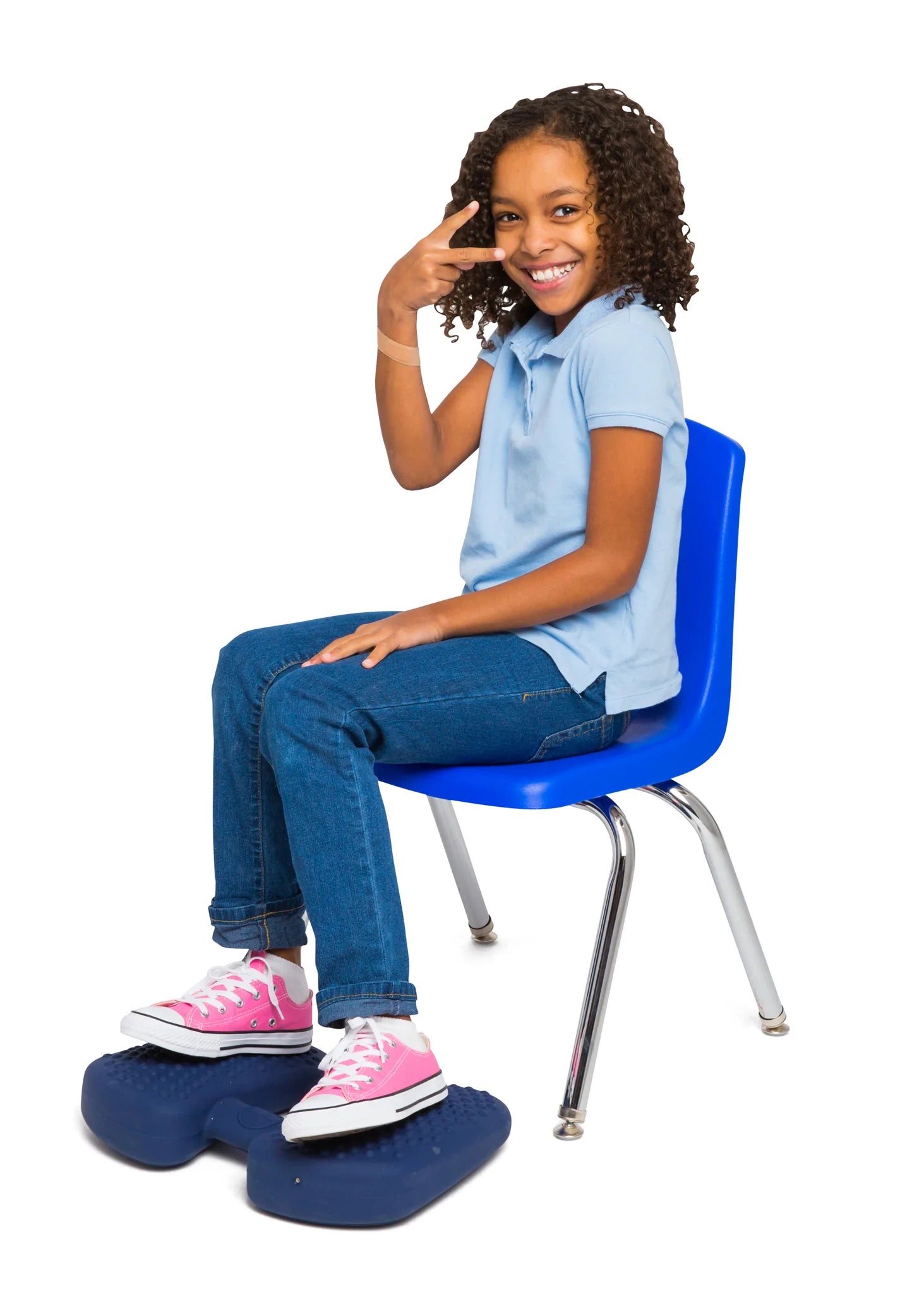 A child with medium skin tone and curly dark brown hair sits on a school chair. Their feet are resting on Wiggle Feet and they are smiling with their hand in a pose in front of their face.
