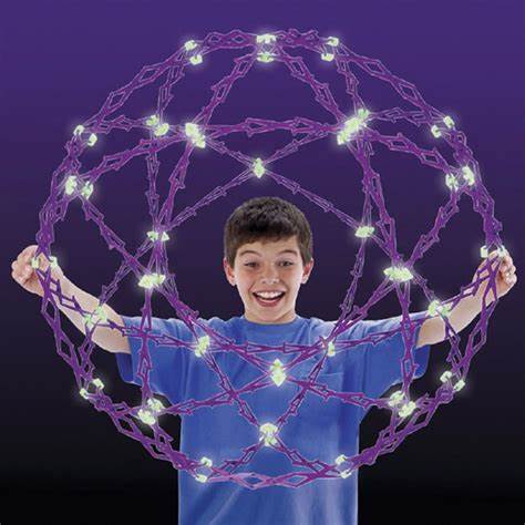A child with light skin tone and short brown hair holds an expanded Hoberman Sphere Glows in front of their chest. Several of the linked pieces are glowing in the dark.