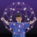 A child with light skin tone and short brown hair holds an expanded Hoberman Sphere Glows in front of their chest. Several of the linked pieces are glowing in the dark.