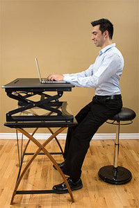 An adult with light skin tone sits up high on an Adult Office Plus Sit-Stand Wobble Chair at a desk with an open laptop.
