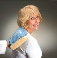 An older adult with light skin tone and shoulder length blonde hair is smiling at the camera. Someone is holding the Vibrator Mitt up to their right shoulder blade.