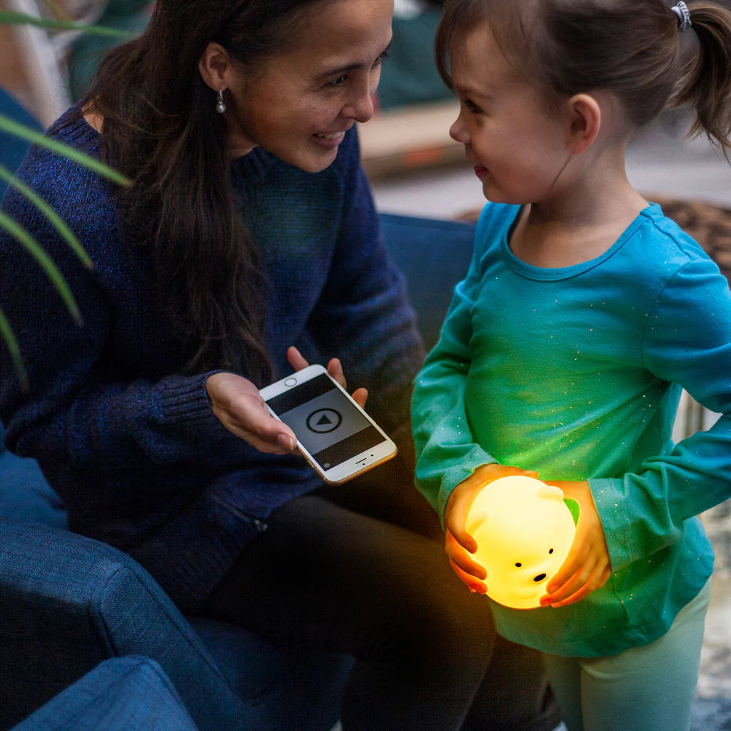 An adult with medium light skin tone and long dark hair is smiling at a child. The adult is holding a phone. The child has light skin tone and brown hair pulled back in a pony tail. They are holding the lit up Bear LED Night Light Bluetooth Speaker.